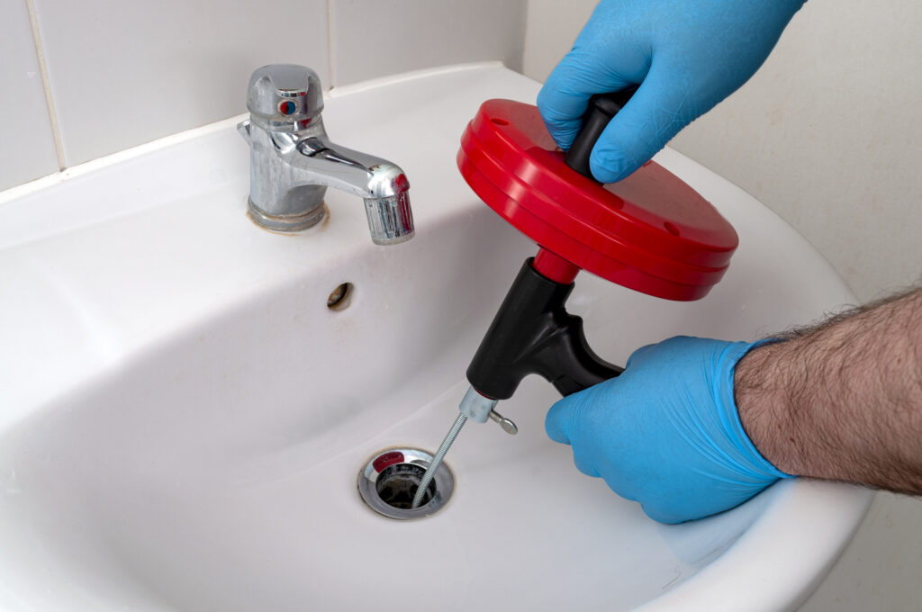 Plumber's hands using a drain snake in a small white bathroom sink drain.